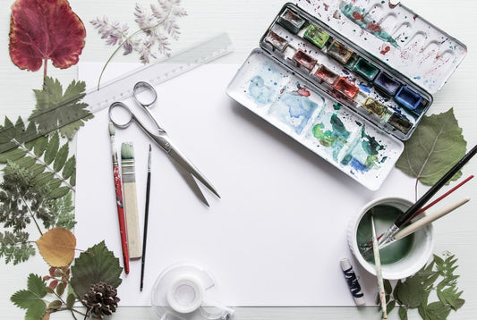 Watercolour paints with paint brushes, pencils, scissors, a ruler and tape on a piece of paper surrounded by a selection of leaves.